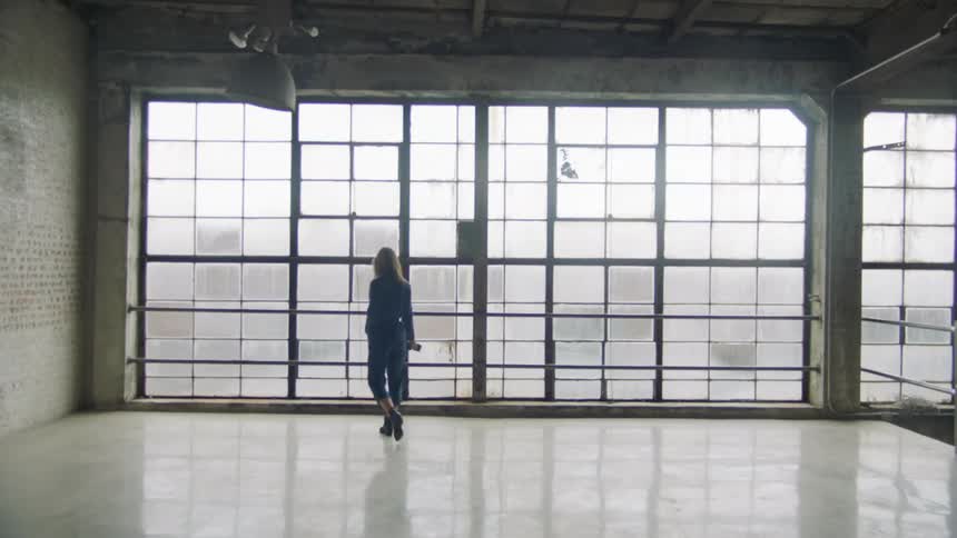 A woman carrying a camera walks in to an empty room and looks out the window
