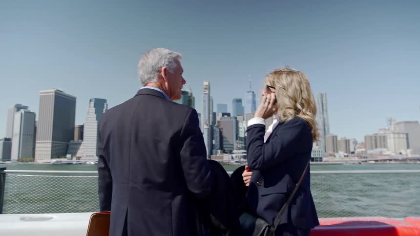 A man and a woman in business attire stop at a fence by a river and talk
