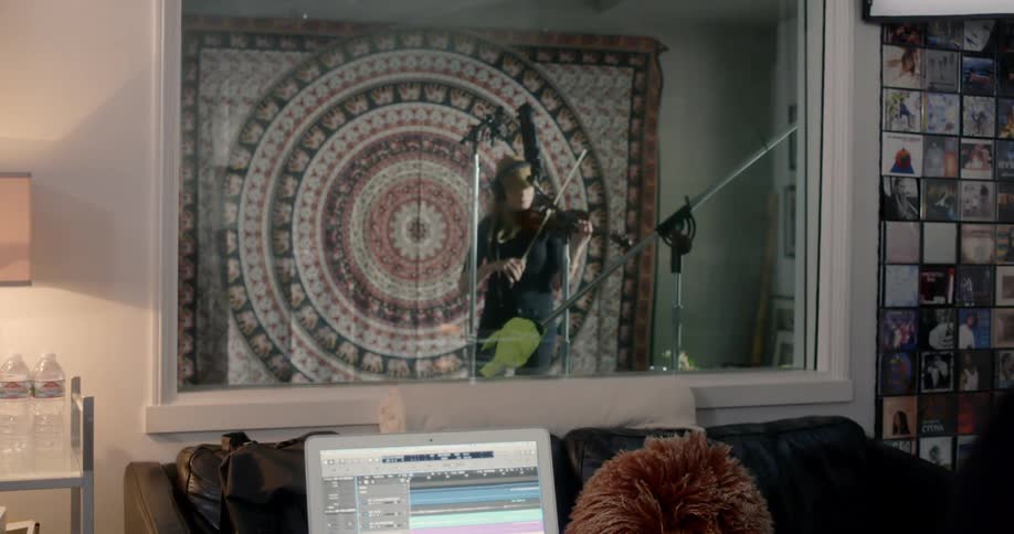 A man in front of a laptop looking at a woman playing the violin in a sound booth