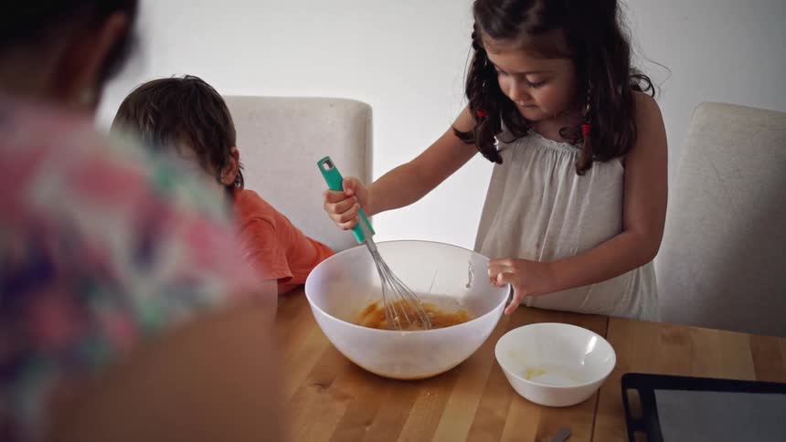 A girl and a boy sitting at a table as the girl mix a bowl and a woman hands the boy a bowl of flour