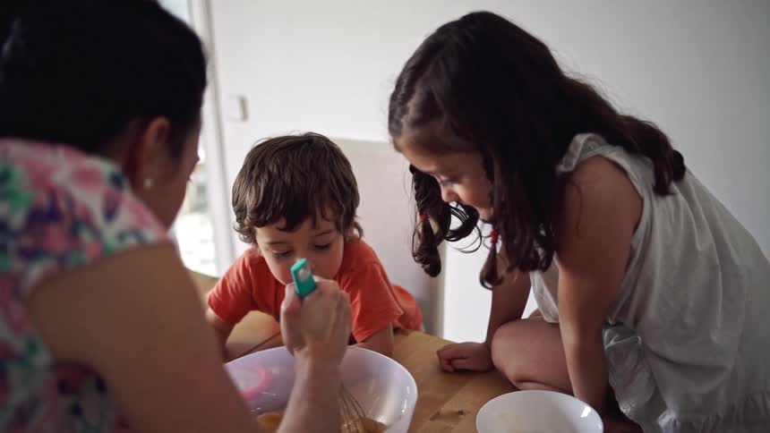 A little girls sits on the wooden kitchen counter while her brother leans against it watching their mom mix batter