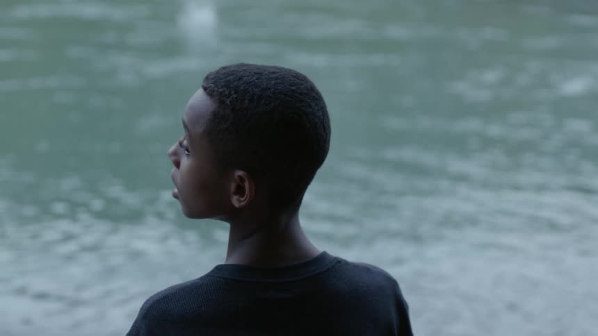 A black boy stands outdoors by a body of water