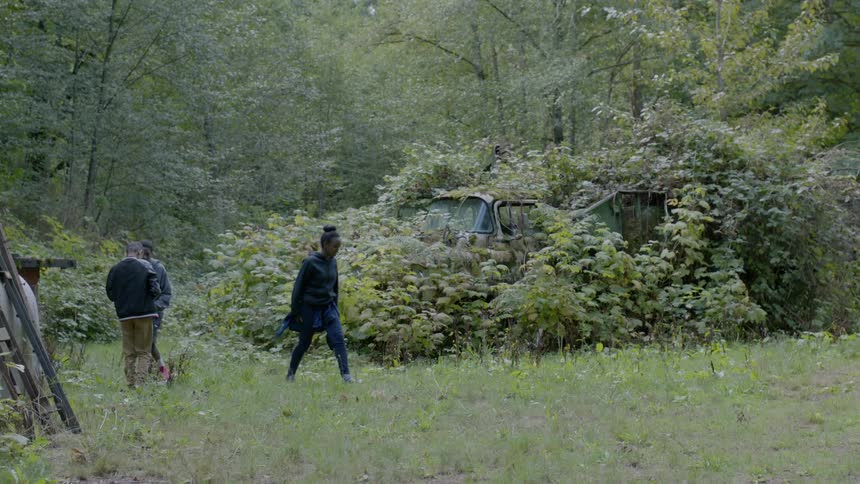 Three black teens walk around a field with old cars overgrown in bushes
