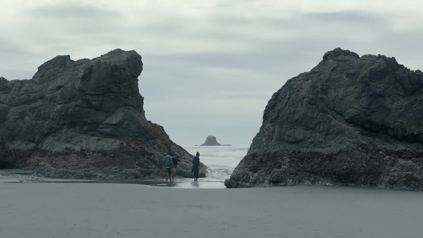 Three young people play at edge of water between two huge boulders on beach