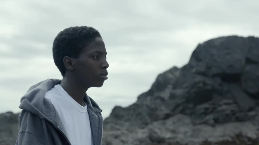 Teenage black boy on rocky beach under grey sky