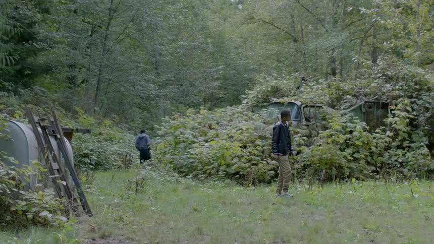 A boy carrying and playing with a stick walks around and looks at an abandoned truck covered in ivy and brush