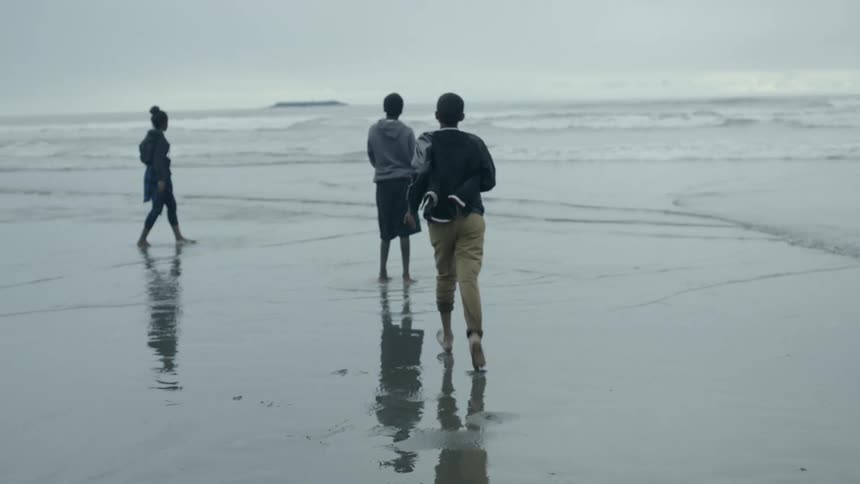 A boy running along a beach to greet another boy and girl who are tossing stones into the ocean