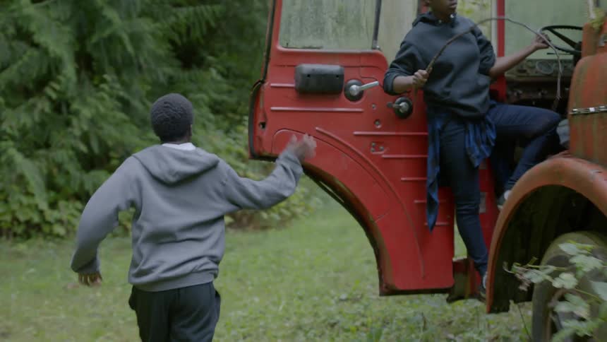 A boy jumping off the back of a dilapidated truck in a woodland and running pass a boy and girl playing with sticks