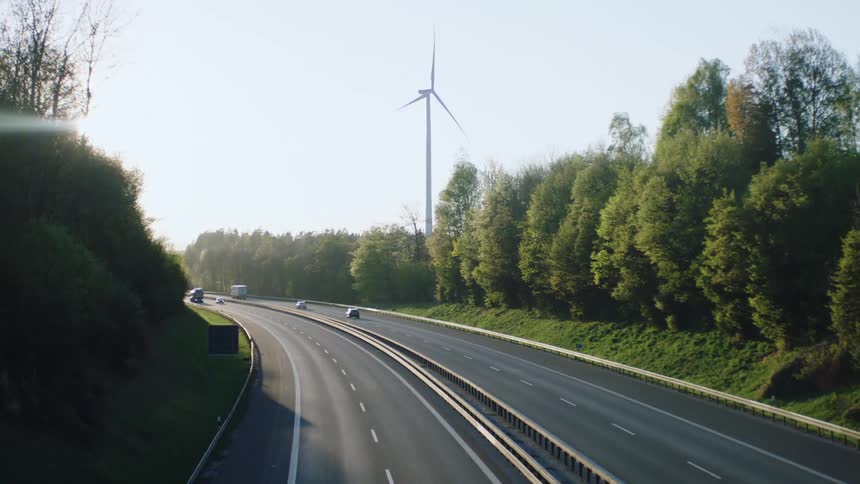 Light traffic on four lane freeway through trees in front of wind turbine