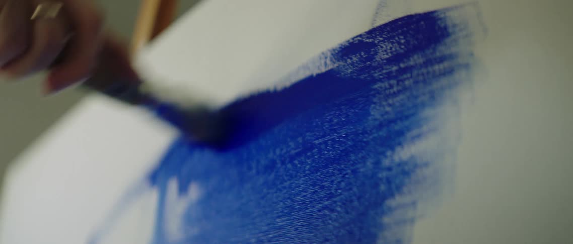 A white woman brushes blue paint on a white canvas