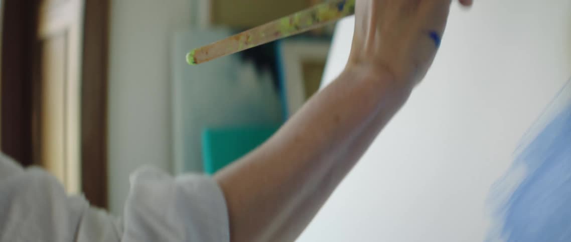 A painter covering a paint brush in yellow paint before raising it to paint on a canvas