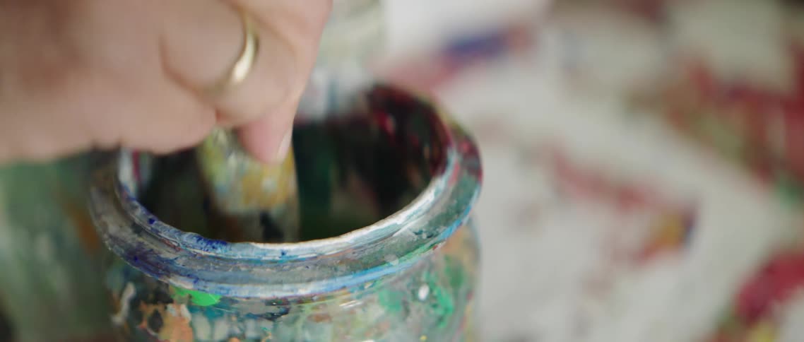 Female places paintbrush covered with blue paint into the water of a glass jar splattered with different colors of dried paint and moves it up and down to clean the paint off of the brush