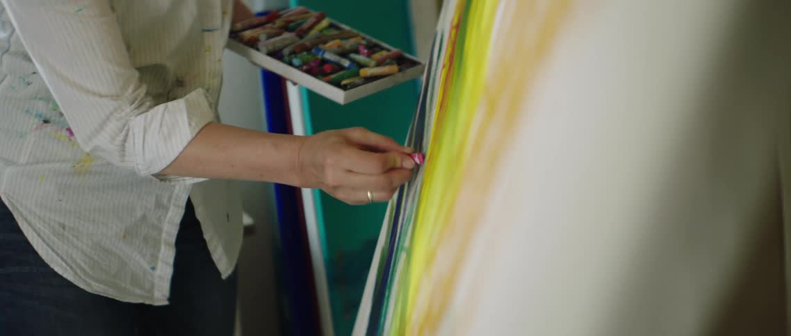 A woman holds a box of artist chalk and uses them on a canvas