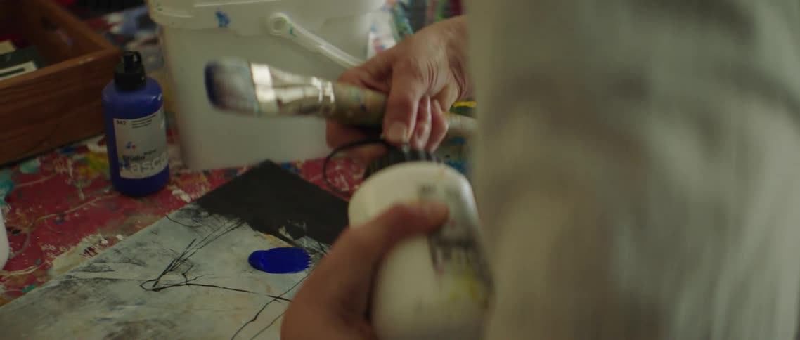 A person picks up a bottle of paint and shakes it before squirting some onto a piece of paper