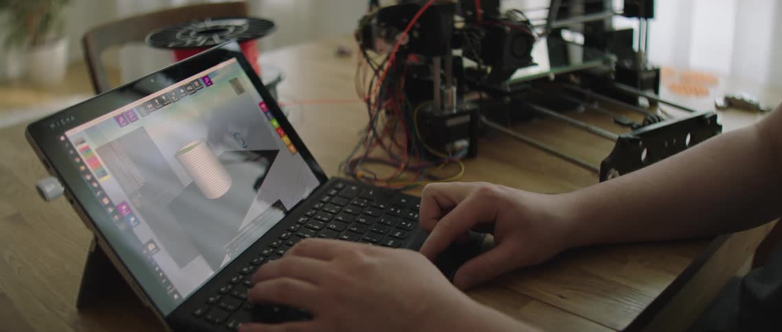 A person sits at a table with a laptop and 3d printer