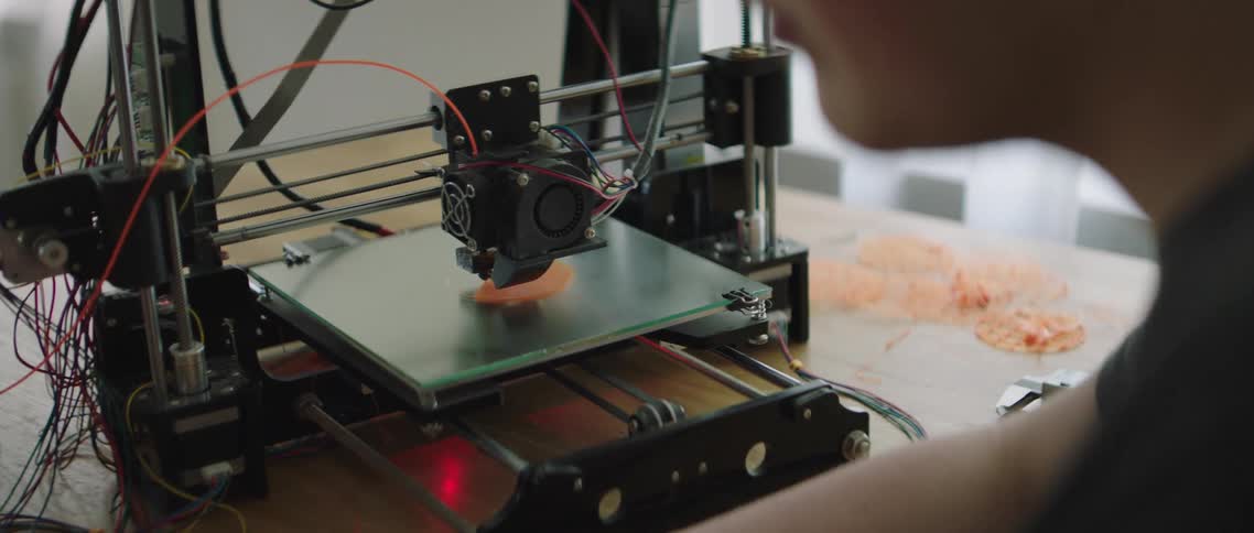 A person sitting next to an a 3d printer as it prints an object
