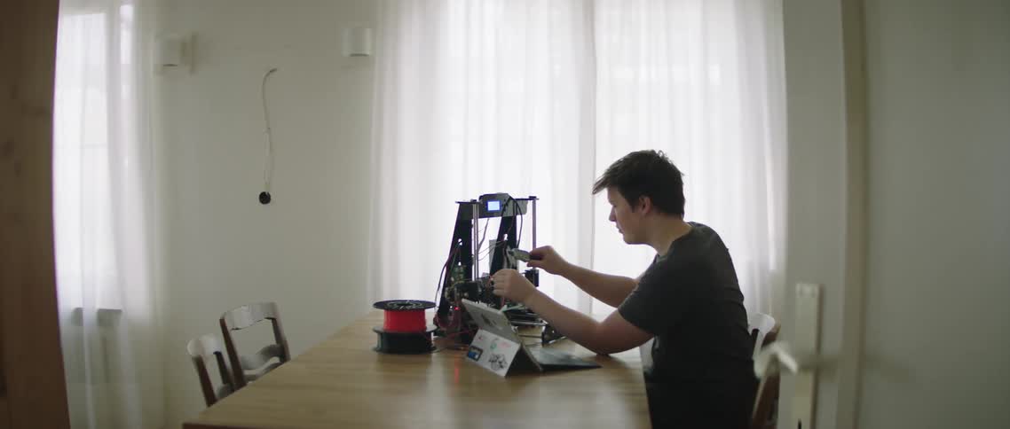 A man sitting at a table works on a 3d printer and his laptop