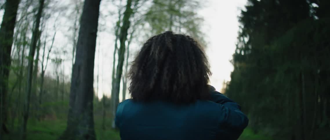 A woman stands in a rural tree filled area with a camera to her face