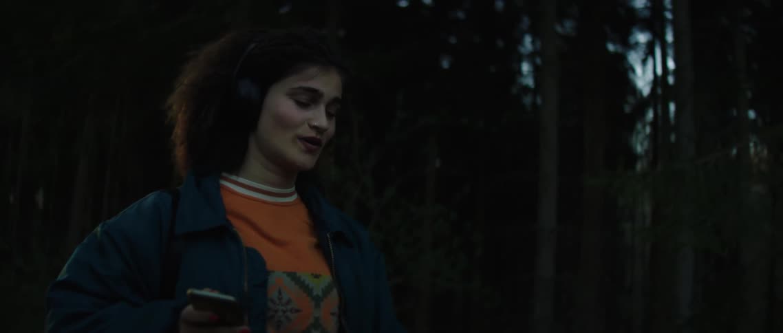 A young woman walking down a road through a woodland while singing along to a song playing in the headphones connected to her smartphone