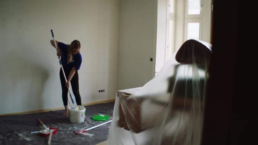 A woman dips a long handle paint roller into a bucket of paint