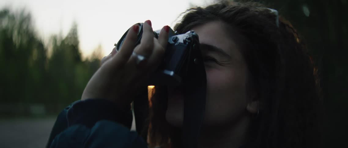 Young woman points camera and takes photo on road through forest at dusk