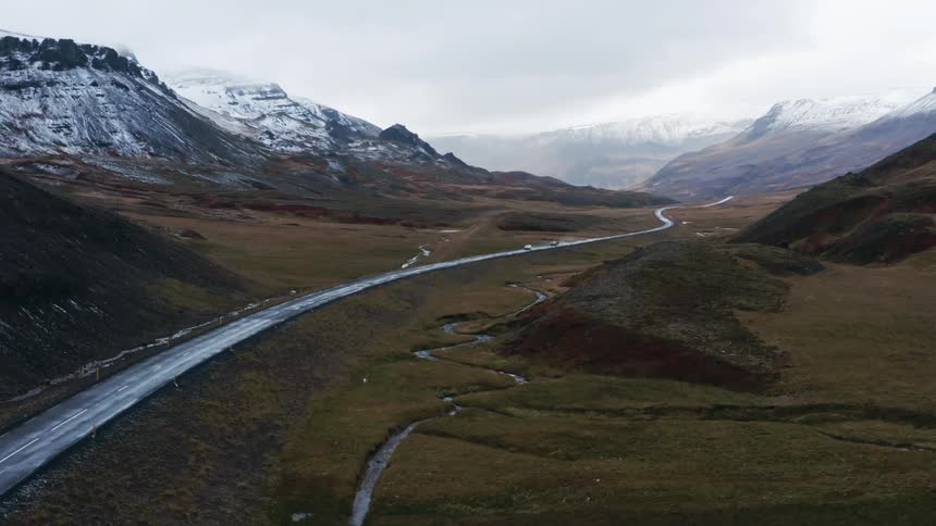 Cars travel on a wet slick road with snow dusted mountains behind and a winding stream beside
