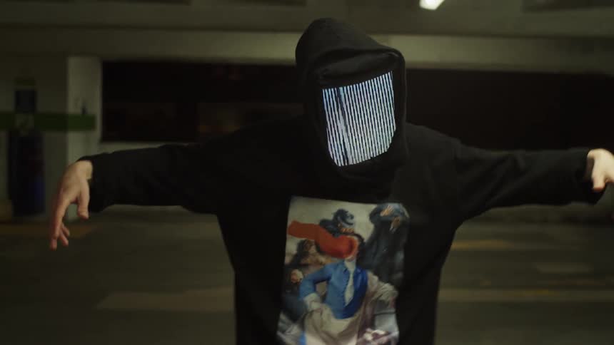A dancer with a screen for a face moves his arms around in a parking garage