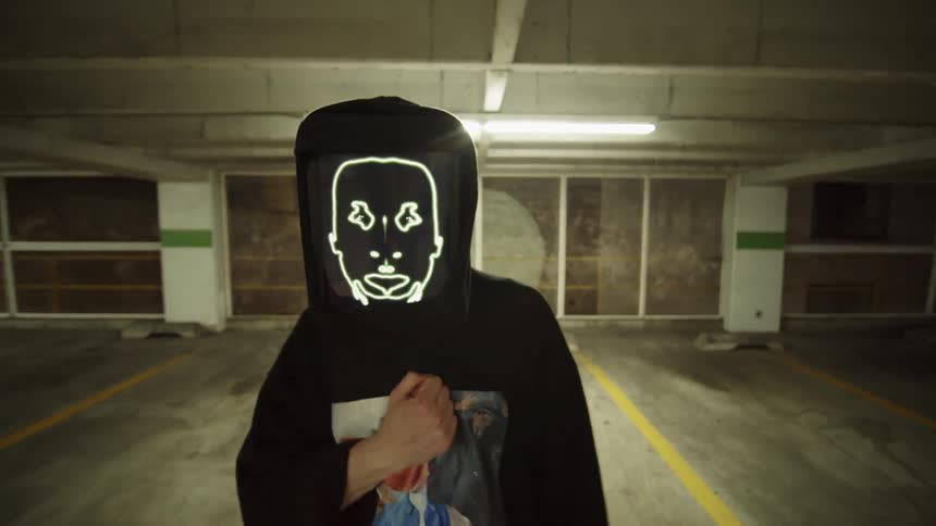 A rapper with a display screen for a face raps while walking through an indoor garage