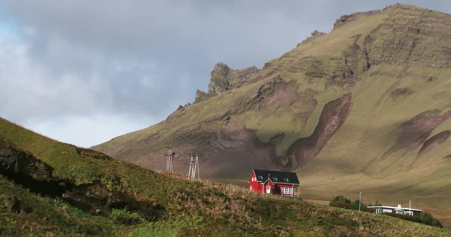 A bright red house sits on the green slope of a hill with a large rocky hill behind