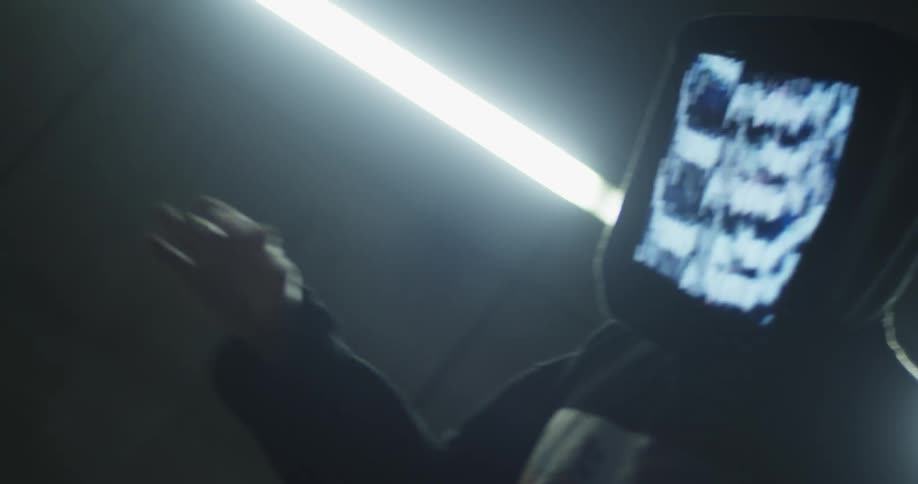 A rapper with a digital screen for a face dances inside a building