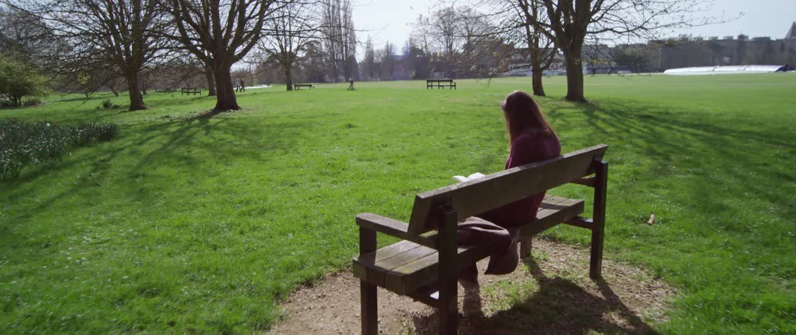 Coming up behind a woman sitting on a park bench reading a book