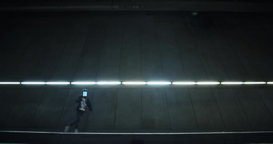 A person wearing a digital display with a hoodie as a mask dances in front of a tall wall