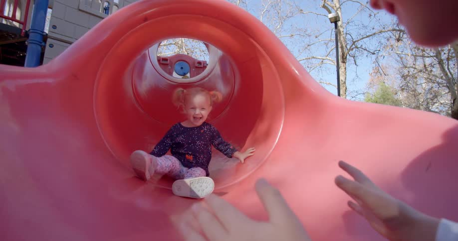 A little girl with pigtails slides down the slide laughing at her waiting mom