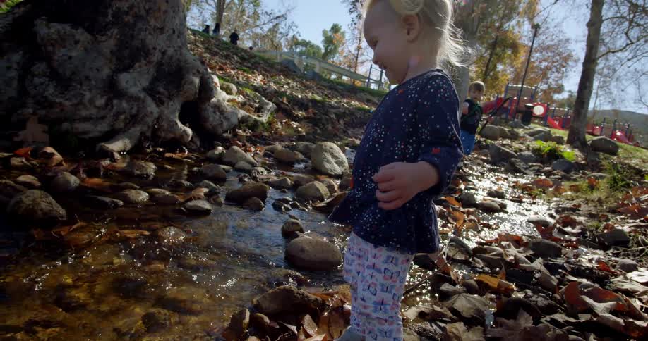 A white female toddler throws a rock in a stream near a playground