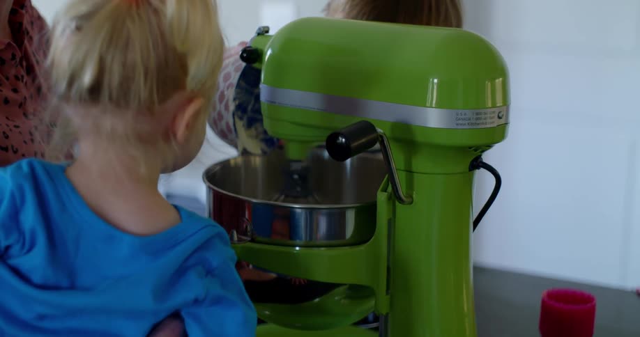 A woman is pouring ingredients into a stand mixers bowl as her grandchildren look on