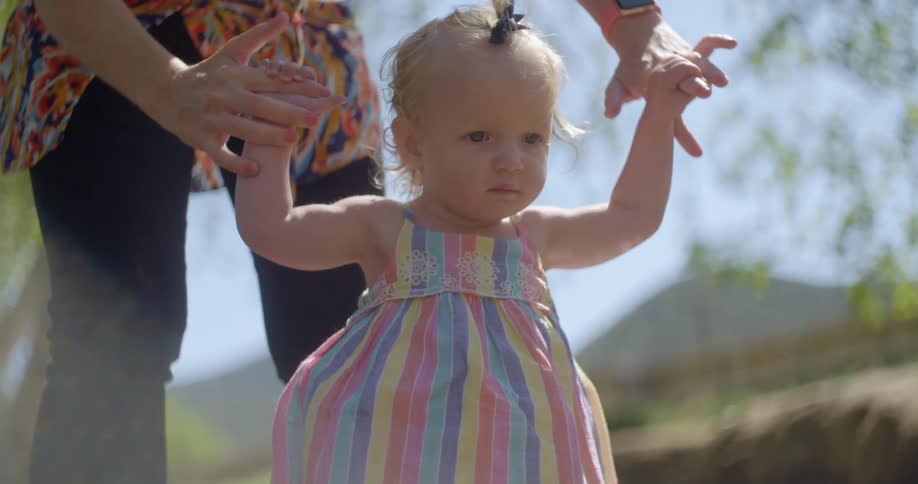 A little girl holds her moms fingers as she stands on her own on a bright sunny day