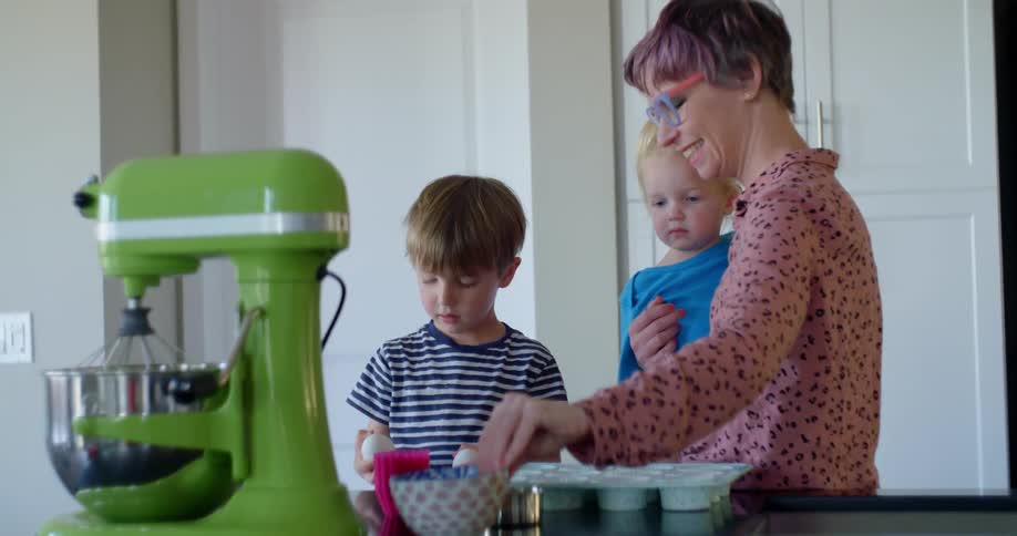Mother holding toddler in kitchen with little boy cracking eggs into cup