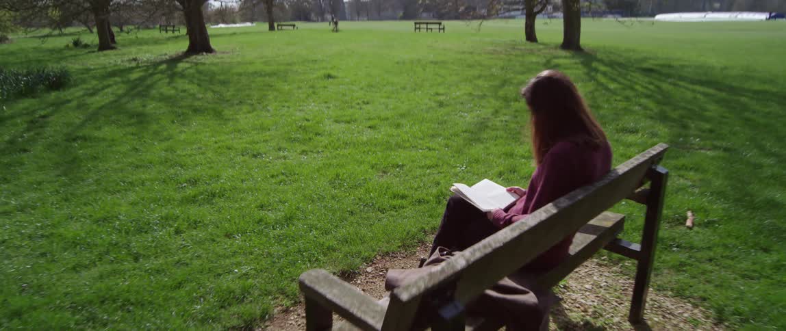 A white woman sits on a bench and reads a book in the park