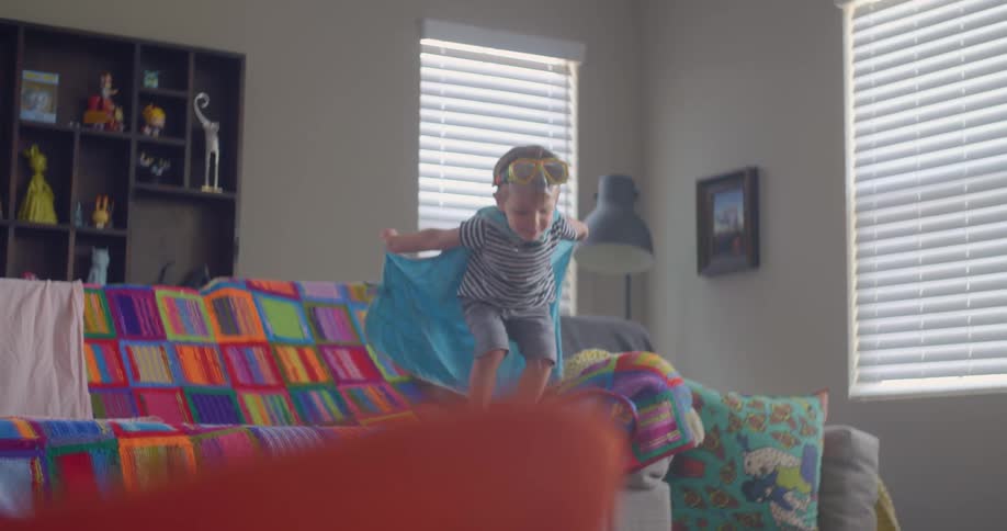 Little boy with blanket cape jumps off of sofa