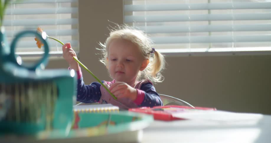 Toddler girl sitting at table smells flower and smiles