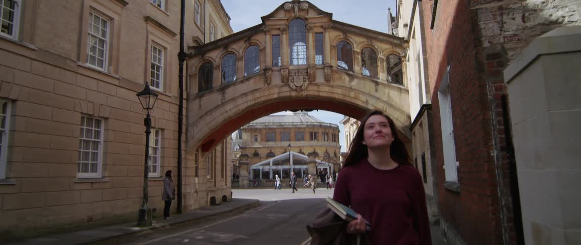 A student walks through a historical campus holding books near Hertford Bridge