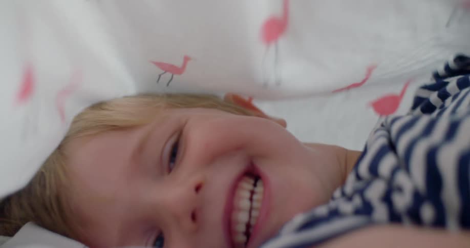 A young boy lies in bed with a sheet over his face and laughs