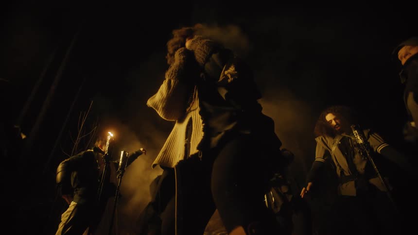 A woman looks around frightened at the group dancing around her by candlelight at night
