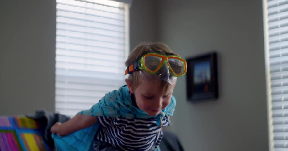 Child wearing a blanket as a cape jumps from a couch in the livingroom of a house