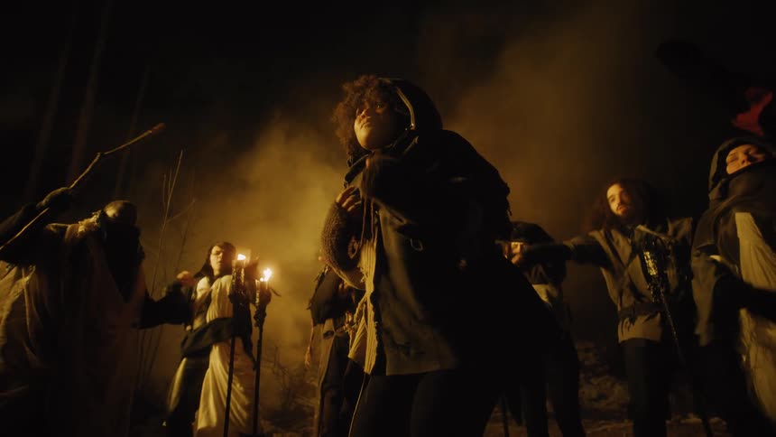Group of people dancing and chanting around woman by candlelight in woods at night