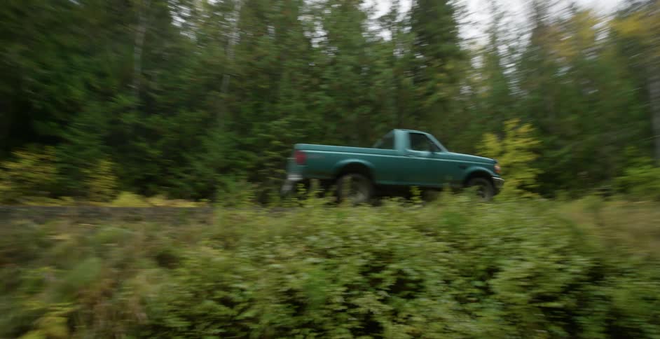 Older model pickup truck drives down rural road through forest