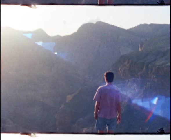 Man wearing baseball cap and sunglasses stands atop mountain at sunset