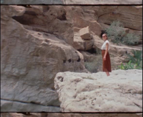 A white woman in a dress stands on an outcropping of rock near a shallow pool