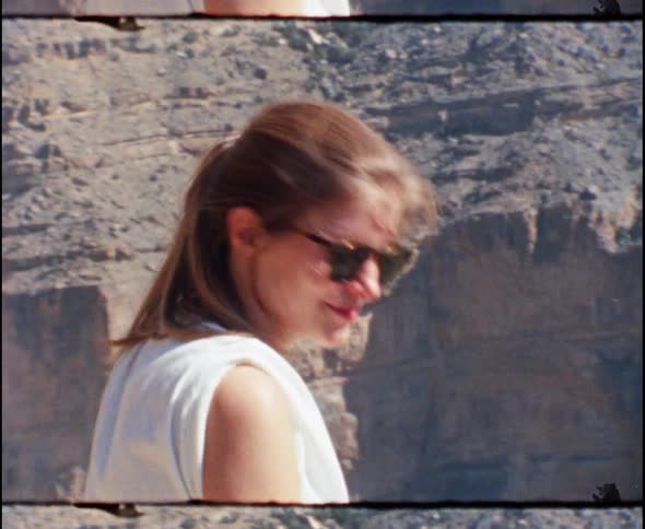 A white woman wearing sunglasses sits on the edge of a large canyon