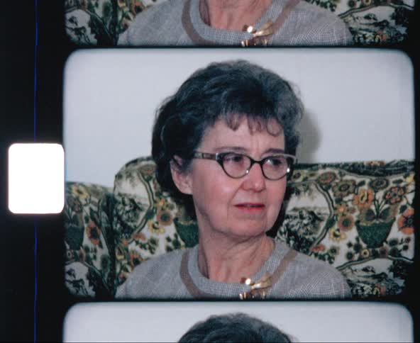 An elderly woman speaking and smiling while gesturing as she sits on a sofa